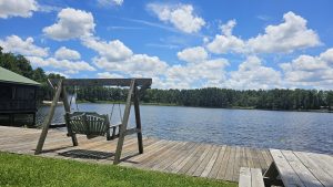Wooden swing on the dock overlooking Lake Como at Lake Como Family Nudist Resort in Florida, surrounded by pine trees and blue skies.