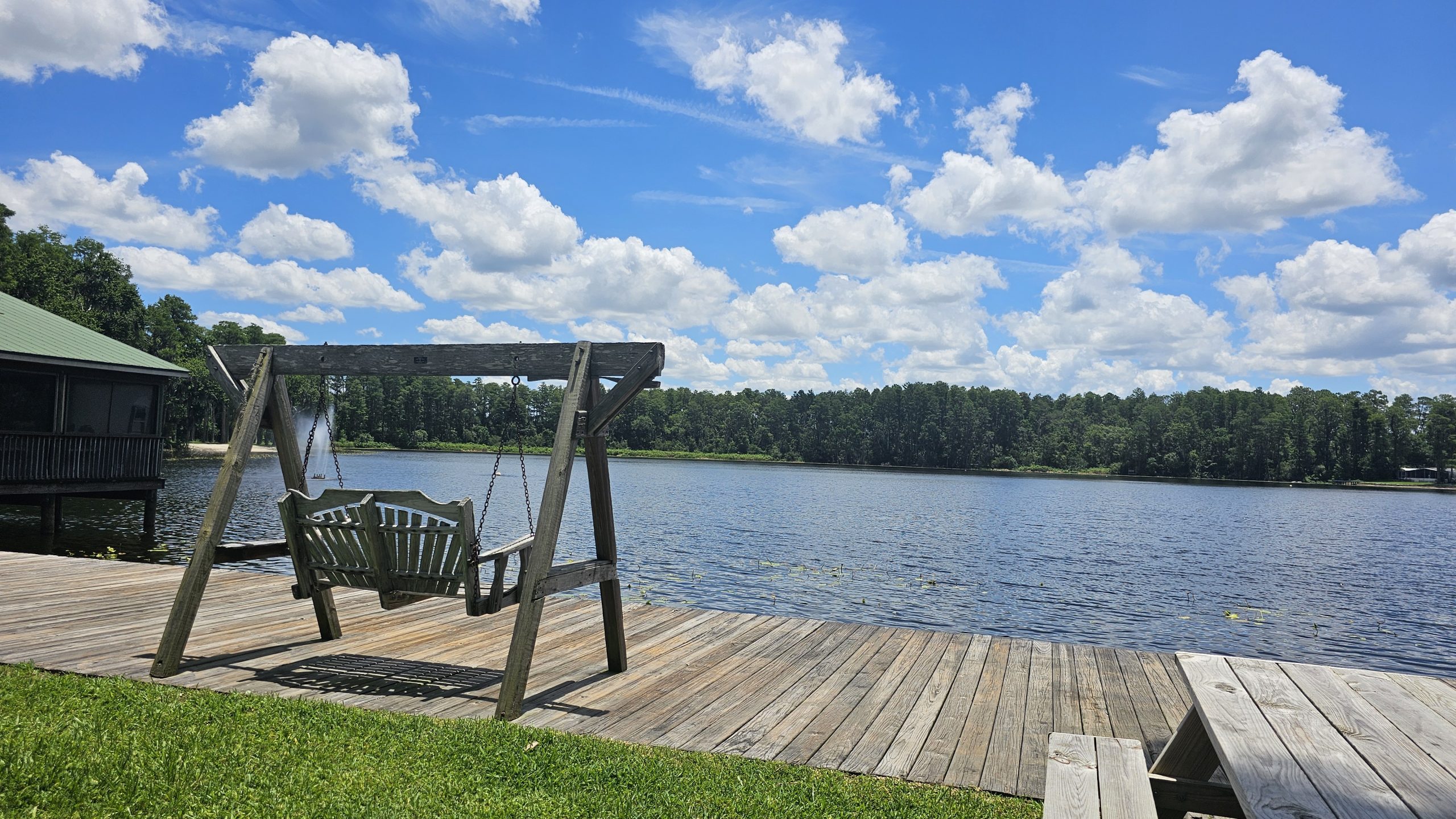 Wooden swing on the dock overlooking Lake Como at Lake Como Family Nudist Resort in Florida, surrounded by pine trees and blue skies.